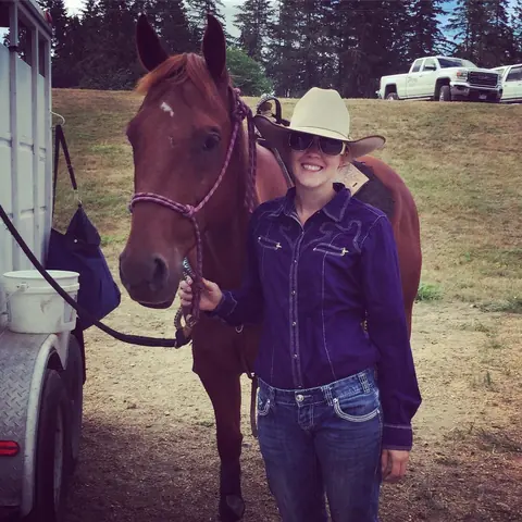 Alyssa in a cowboy hat standing with a bay horse beside a trailer